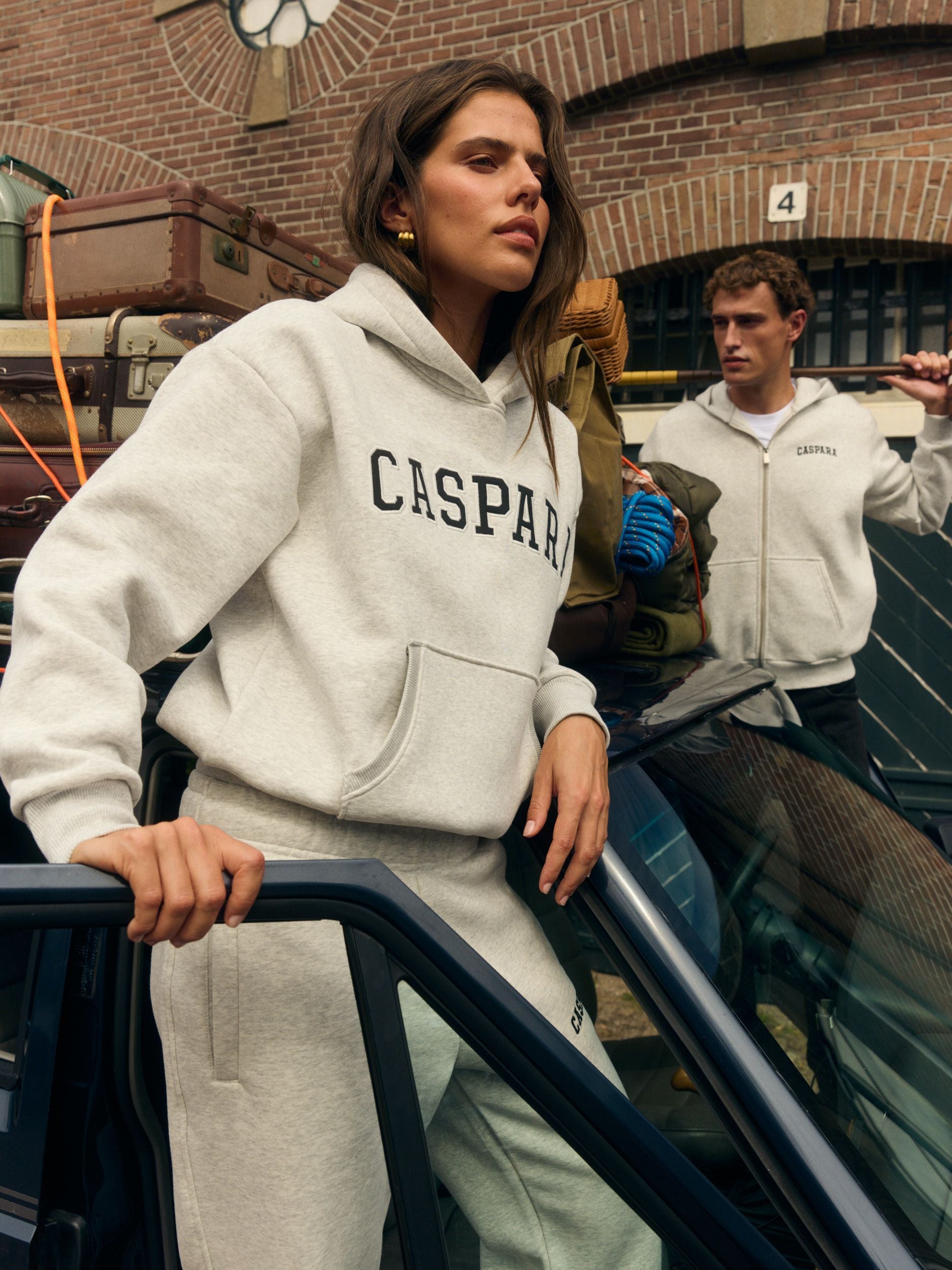 Woman wearing a grey melange hoodie and sweatpants leaning against a car packed with luggage.